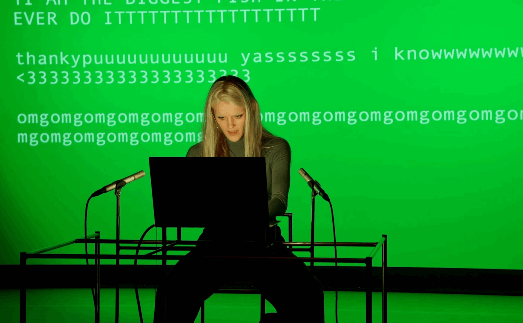 Een persoon zit achter een laptop met groene achtergrond, omringd door microfoons, en lijkt enthousiast.