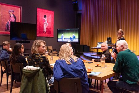 Groep mensen zit rond een tafel in een zaal met kunst aan de muur, tijdens een bijeenkomst of gesprek.