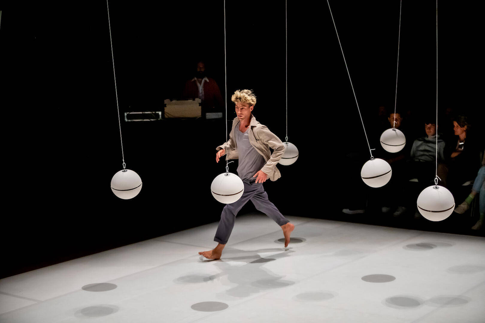 Een danser in moderne kleding beweegt tussen hangende lichten op een podium, met een publiek op de achtergrond.