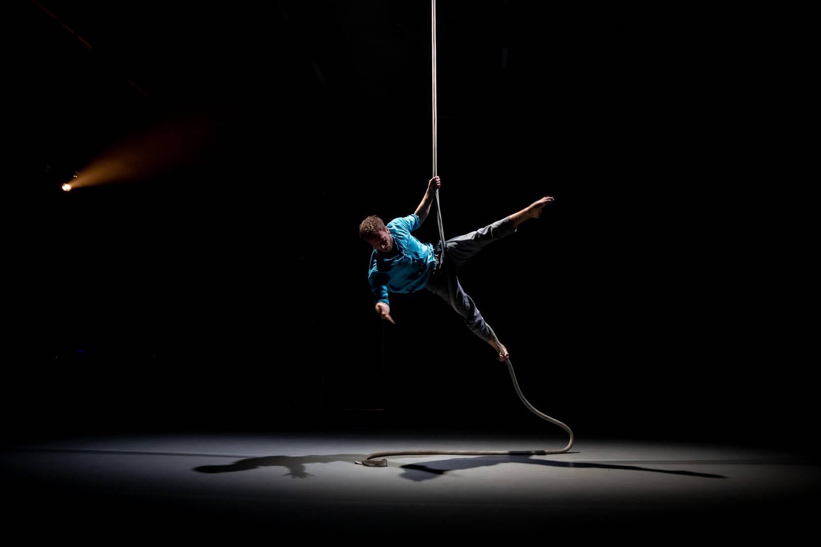 Een artiest voert een acrobatische act uit aan een touw in een donkere ruimte met spotlichten.