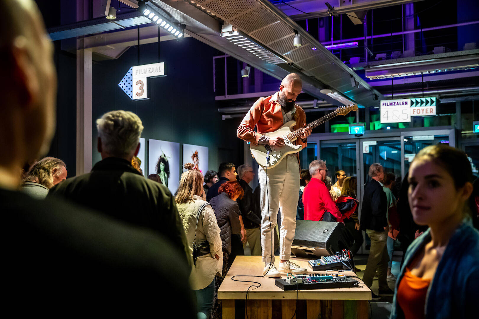 Een muzikant speelt gitaar op een verhoging, terwijl publiek in een moderne ruimte toekijkt.