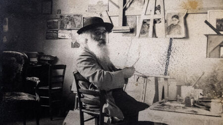 Oude zwart-witfoto van een oudere man met een baard, zittend op een stoel in een atelier vol kunstwerken.