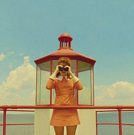Een vrouw in een oranje jurk kijkt door verrekijkers vanuit een lantaarnhuisje aan het water onder een blauwe lucht.