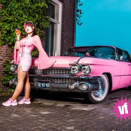 Vrouw in roze outfit poseert met een roze Cadillac voor een gevel met groene klimop.