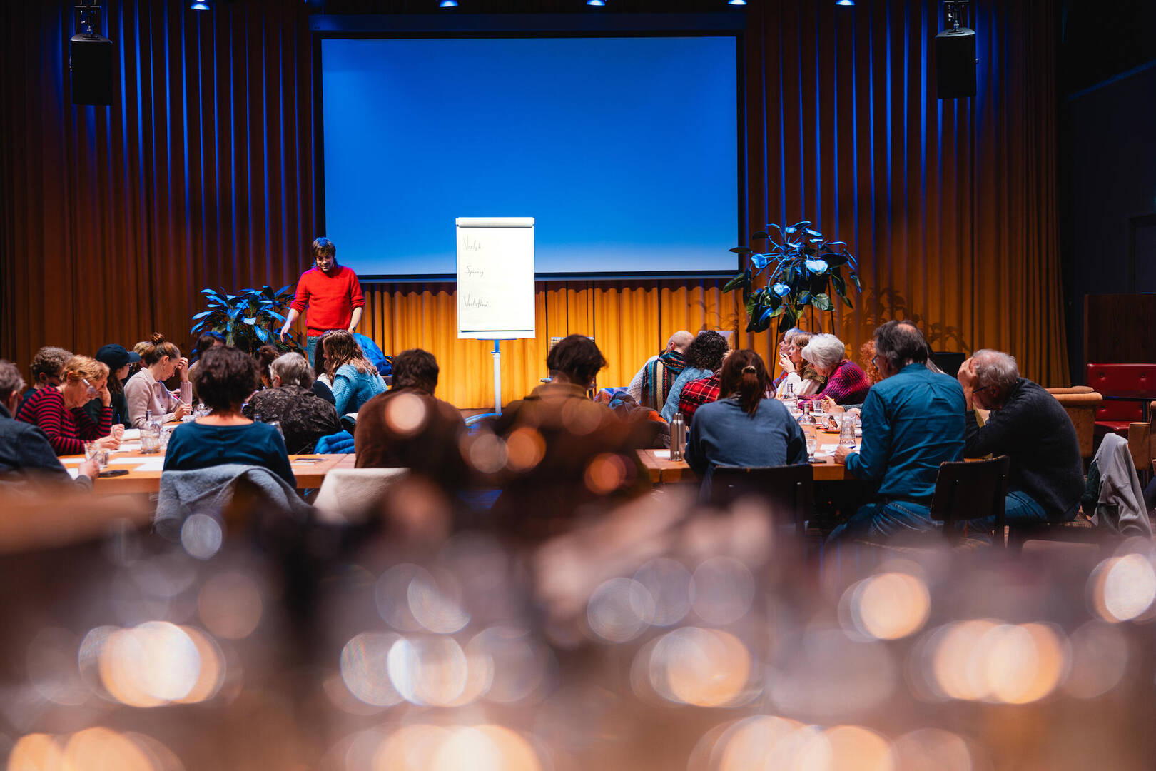Een groep mensen zit aan een tafel in een zaal, terwijl een spreker voor een presentatiescherm staat.