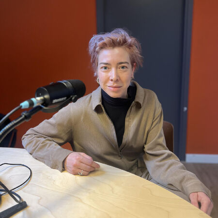 Persoon met lichtroze haar zit aan een tafel met een microfoon in een studio-achtige omgeving.