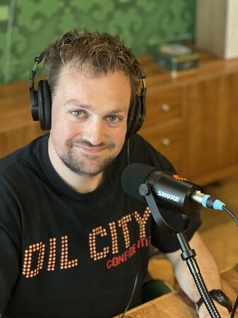 Man met headphones en microfoon, glimlachend, draagt een t-shirt met "DIL CITY" op een groene achtergrond.