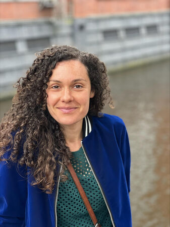 Vrouw met krullend haar in een blauwe jas, staand aan een waterkant met een historische achtergrond.