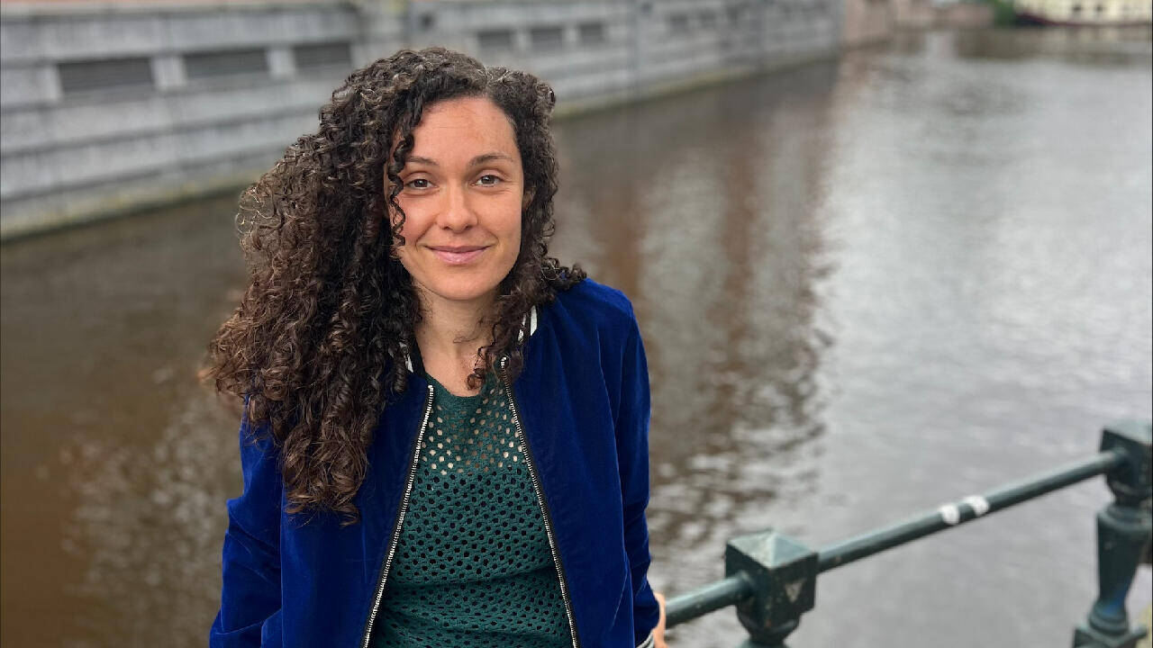 Vrouw met krullend haar in een blauwe jas staat aan een waterkant met een stadse achtergrond.