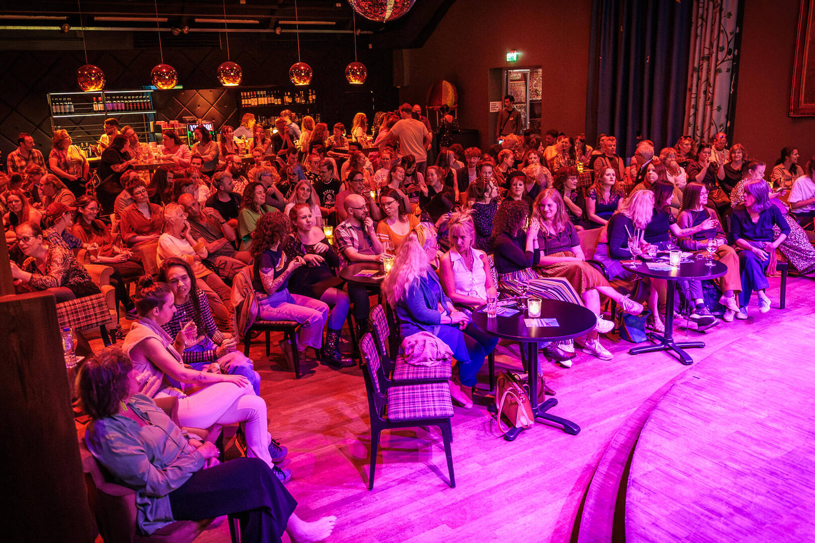 Een volle zaal met publiek dat geniet van een voorstelling, met tafels en een podium zichtbaar.