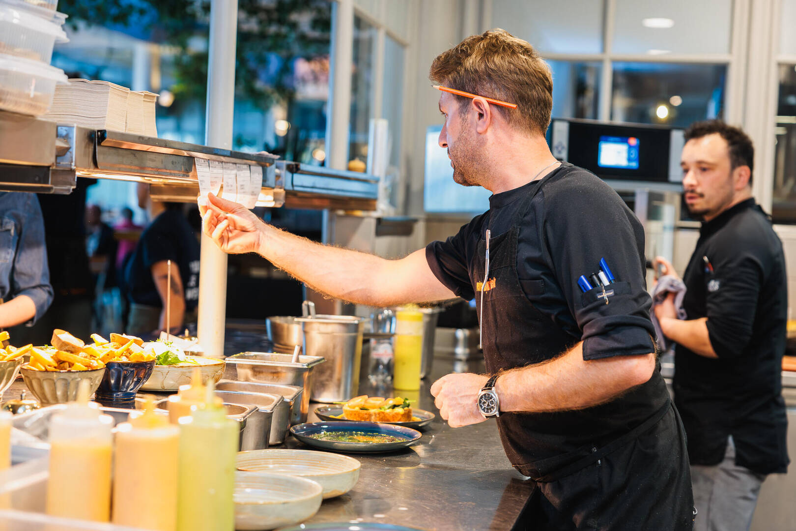 Kok in restaurant bereidt gerechten, terwijl een collega toekijkt. Kleurrijke sauzen en ingrediënten zijn zichtbaar.