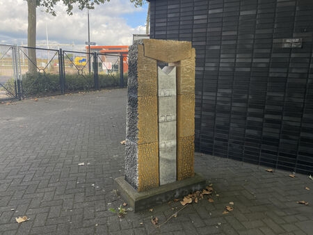 Een rechte, moderne zuil met een gouden en zilveren afwerking, geplaatst op een grindpad naast een zwarte muur.