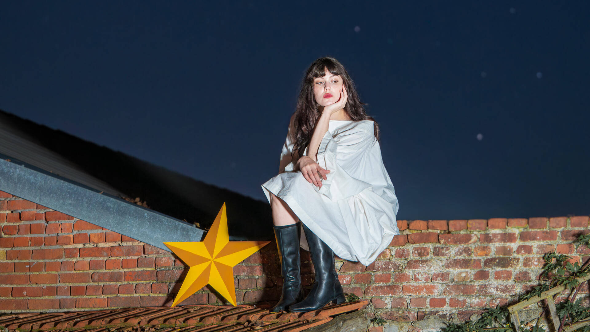 Vrouw in witte jurk en zwarte laarzen, zittend op een muur met een gele ster op de achtergrond onder een sterrenhemel.