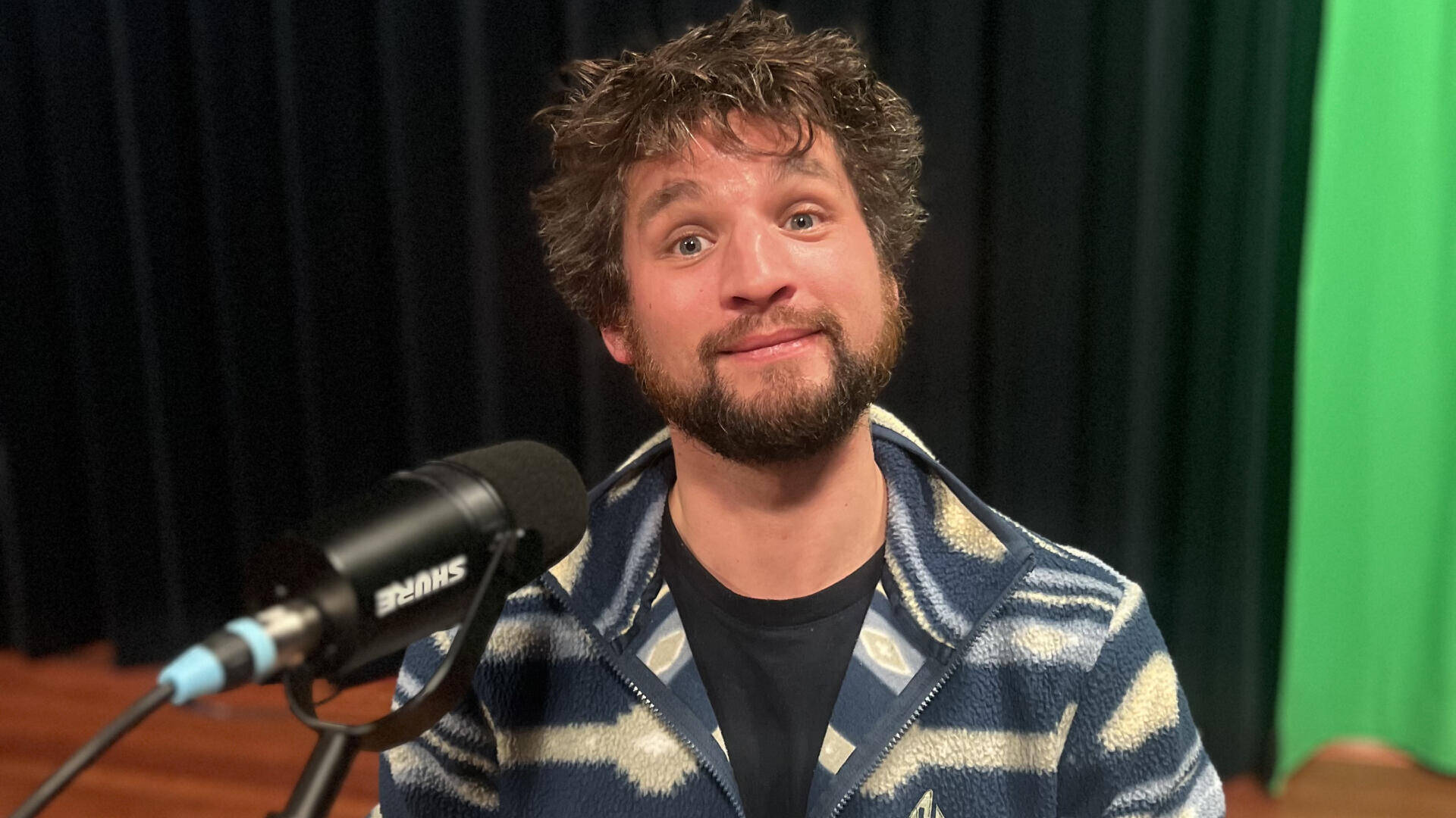 Persoon met krullend haar en een gestreepte trui, lachend voor een microphone tegen een groene achtergrond.