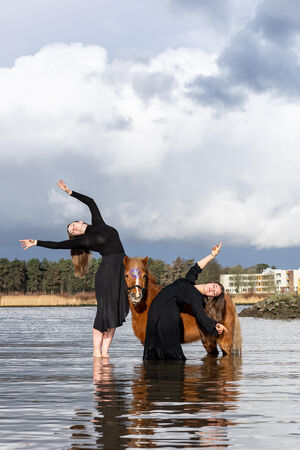 Twee vrouwen in zwarte jurken dansen in ondiep water bij een paard met een bos en gebouwen op de achtergrond.