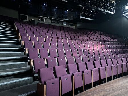 Een lege theaterzaal met paarse stoelen en een trap naar het podium, verlicht door een zacht licht.