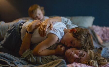 Gezin ligt samen op bed, met een kind dat knuffelt en een ander kind op de achtergrond. Warme, gezellige sfeer.