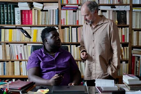 Twee mannen in een bibliotheek, één zittend achter een bureau, de ander staat naast hem en spreekt met hem.