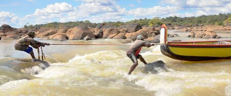 Twee surfers trekken een kano over golven in een brede rivier onder een bewolkte lucht.