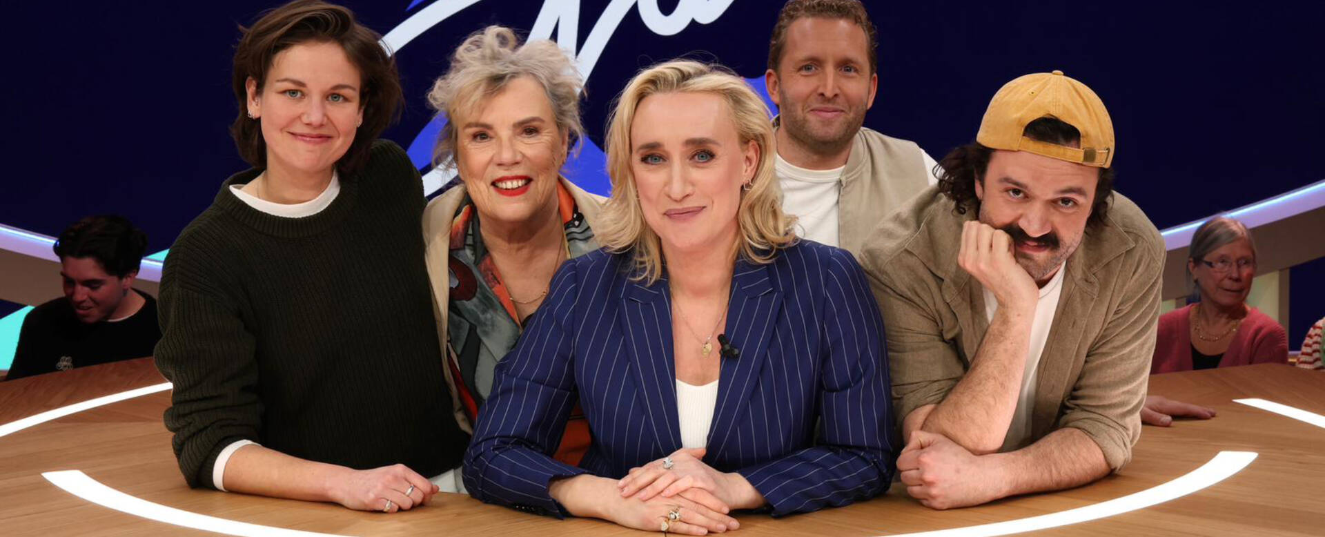 Groep van vijf mensen, waaronder vrouwen en mannen, zittend aan een ronde tafel met op de achtergrond een logo.