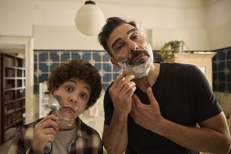 Een man en een jongen staan in een badkamer en scheren zichzelf met scheermesjes. Beide kijken in de camera.
