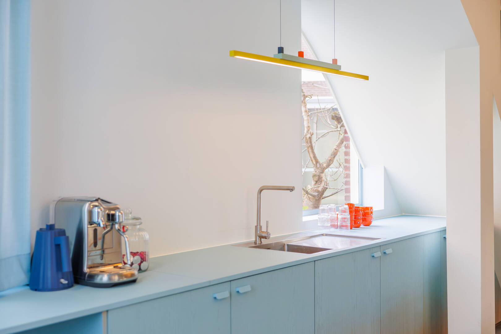 Een moderne keuken met een blauwe kastenwand, een glazen spoelbak, een waterkoker en kleurrijke bekers op het aanrecht.