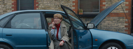 Vrouw in een beige trenchcoat zit naast een blauwe auto met open deur, terwijl ze op haar telefoon kijkt.