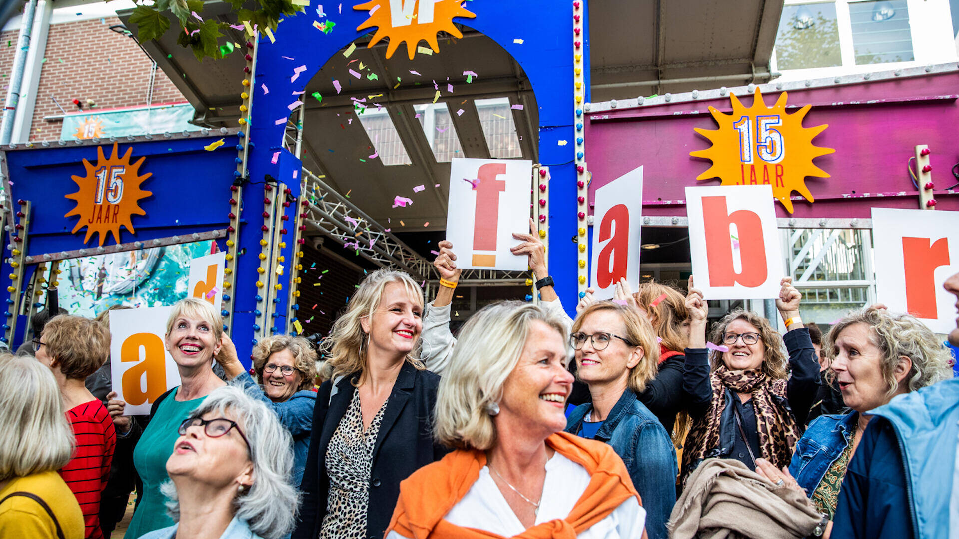 Vrouwengroep staat voor kleurrijke poort Verkadefabriek, houdt borden omhoog met letters, viert feestelijke gelegenheid.