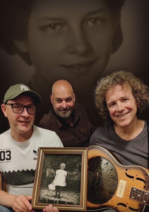 Drie mannen poseren met een foto van een meisje, met een zwart-wit portret op de achtergrond.
