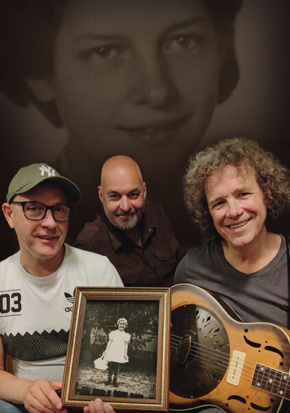 Drie mannen poseren met een foto van een meisje, met een zwart-wit portret op de achtergrond.