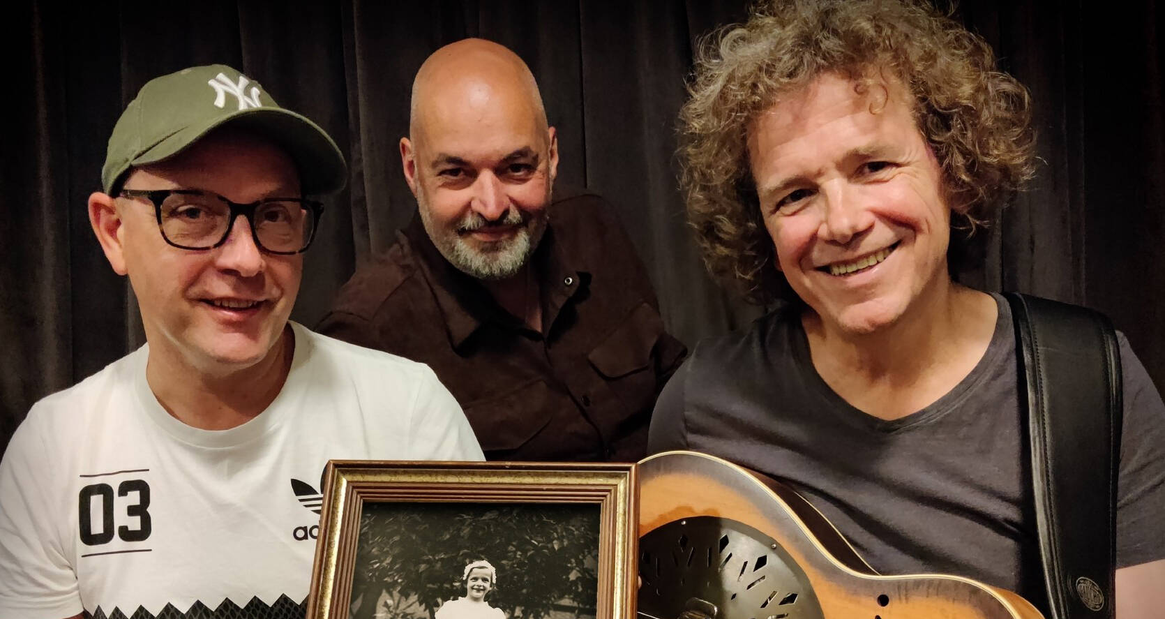 Drie mannen poseren met een oud fotoportret in een fotolijst, lachend voor een donkerbruine achtergrond.