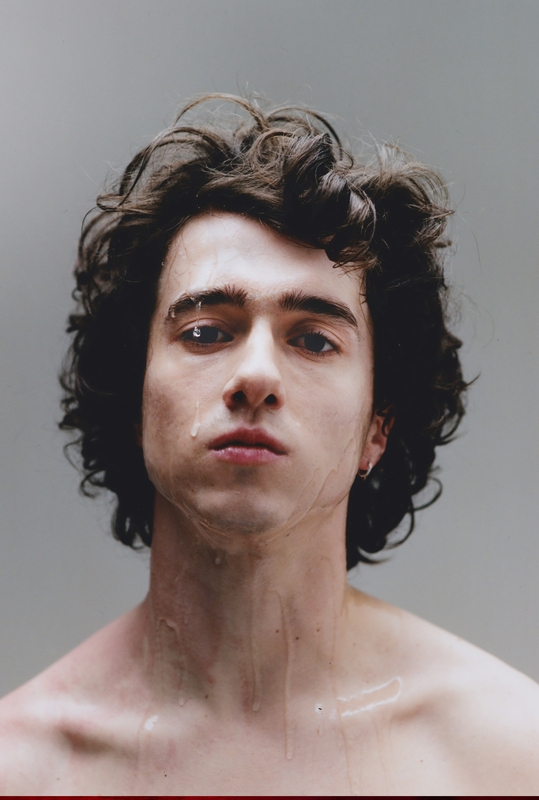 Portret van een jonge man met krullend haar, een neutrale uitdrukking en zonder shirt, met een effen achtergrond.