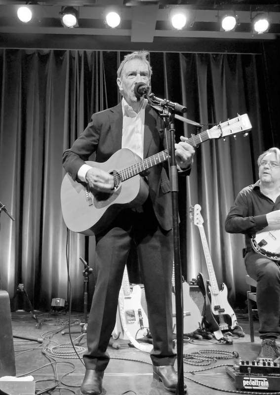 Zwart-wit afbeelding van een man in een pak die gitaar speelt en zingt op een podium met verlichting.