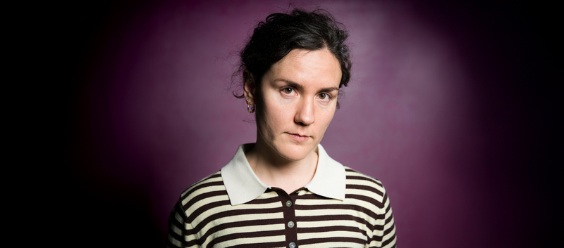 Portret van een vrouw met een neutrale uitdrukking, gekleed in een gestreepte trui, voor een paarse achtergrond.