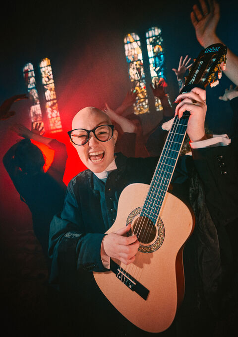 Een bald jongeman met een gitaar, omringd door enthousiaste personen in een sfeervolle omgeving met glas-in-loodramen.