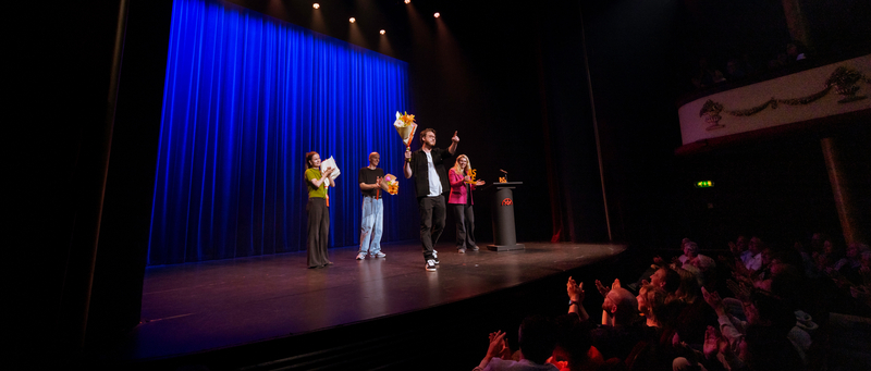 Een groep performers staat op een podium met blauwe gordijnen, terwijl het publiek voor hen applaudeert.