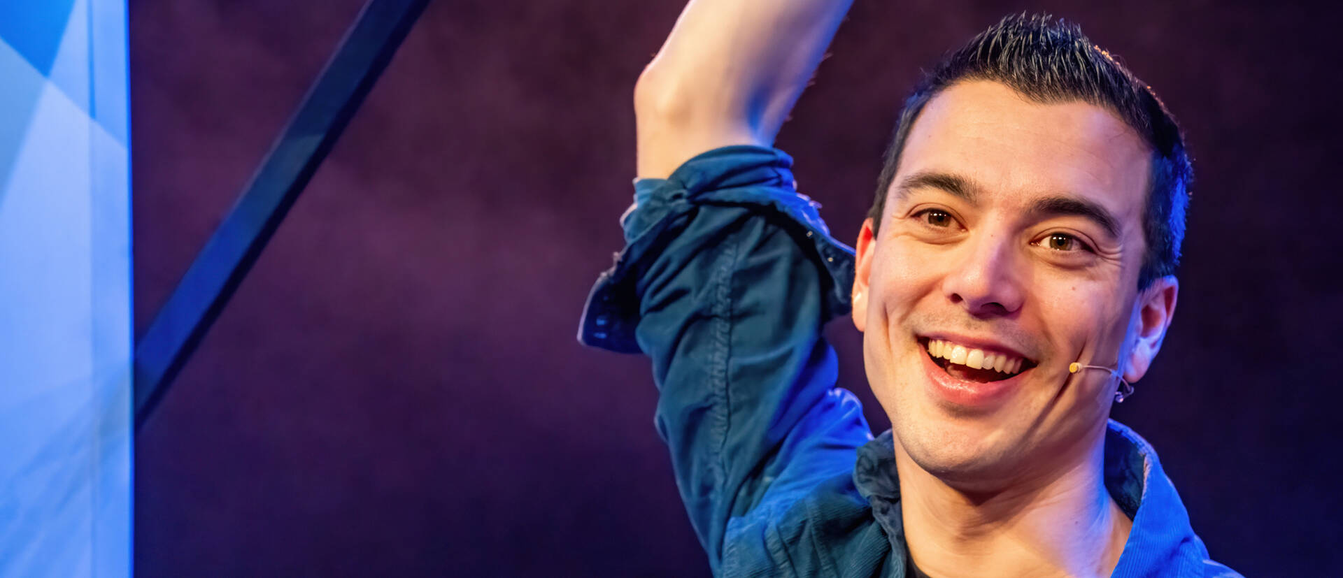 Man met een glimlach die zijn hand omhoog houdt, op een podium met een kleurrijke achtergrond.