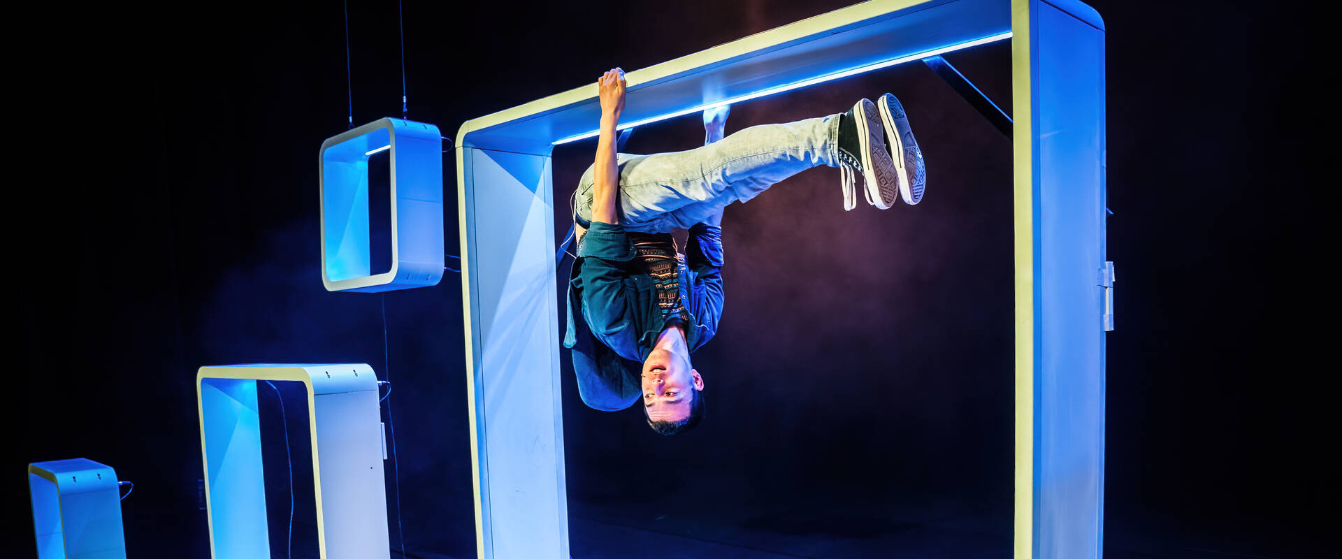 Een acrobatische performer hangt ondersteboven in een lichtblauwe structuur op een podium met kleurrijke belichting.