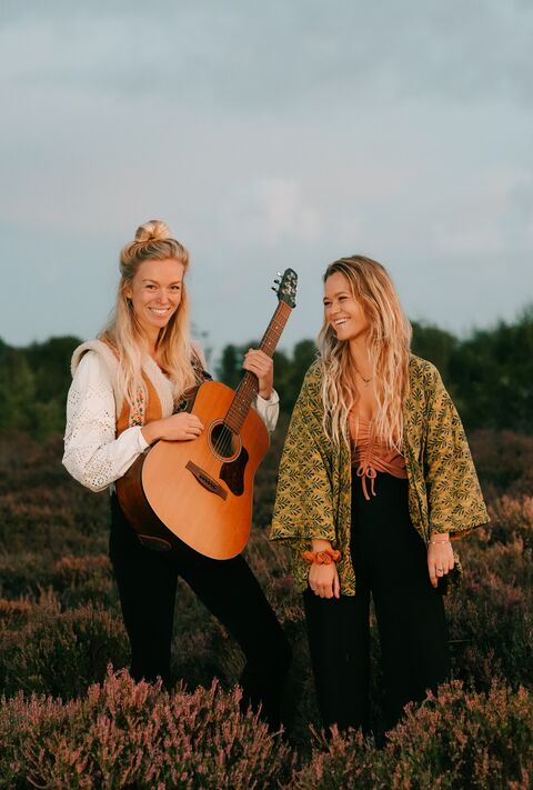 Twee vrouwelijke muzikanten staan op een veld, de ene houdt een gitaar vast en ze dragen kleurrijke outfits.