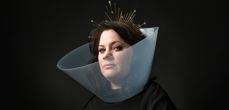 Portret van een vrouw met een geometrische halskraag en een aureool van stijlen, tegen een donkere achtergrond.