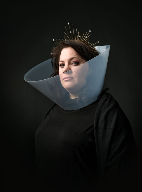 Vrouw in zwart met een opvallende kraag en een kroon van takjes, tegen een donkere achtergrond.