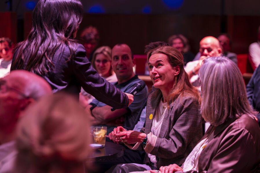 Een vrouw in een zaal ontvangt iets van een andere vrouw, terwijl het publiek zit en kijkt.