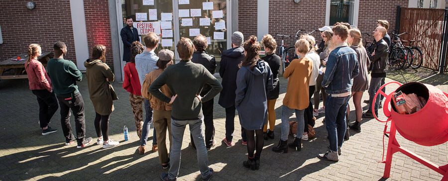 Groep mensen in diverse kleding staat buiten, luisterend naar een spreker voor een muur met affiches.