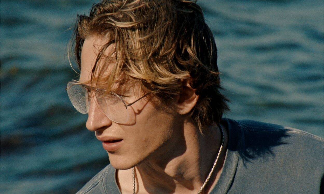 Een jongeman met een casual uitstraling en een licht getinte zonnebril, kijkend naar de wateroppervlakte.