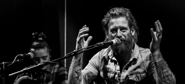 Zwart-wit afbeelding van een muzikant met een baard die in een microfoon spreekt, terwijl een andere muzikant op de achtergrond speelt.