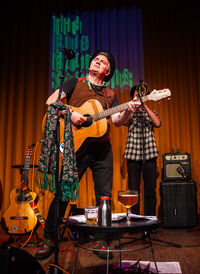 Man met gitaar op podium, terwijl een tweede persoon op de achtergrond staat; een tafel met drank staat voor hen.