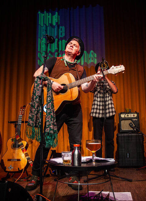 Man met gitaar op podium, terwijl een tweede persoon op de achtergrond staat; een tafel met drank staat voor hen.