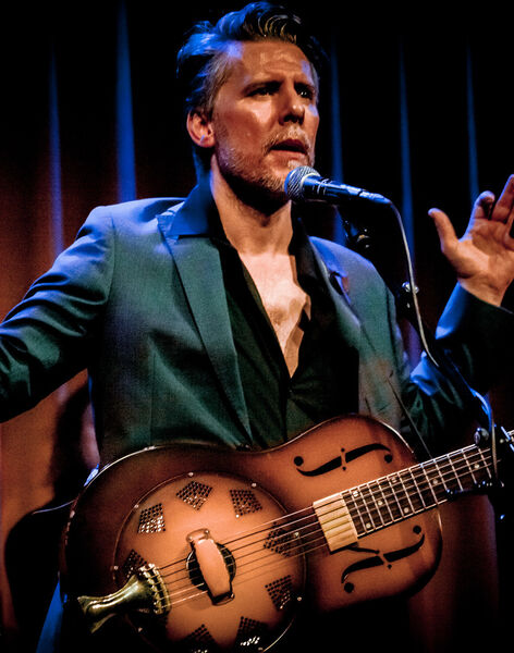 Een man in een blauw kostuum zingt met een gitaar in zijn hand tijdens een live optreden op het podium.