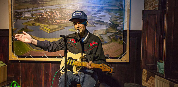 Man speelt gitaar en zingt in een sfeervolle omgeving met een achtergrond van een landschapsschilderij.