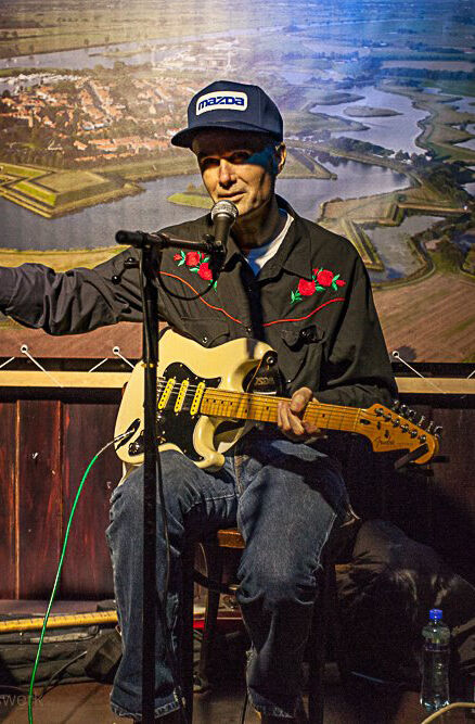 Man met gitaar zingt op een podium, gekleed in een zwart jasje met bloemen en een pet, met een achtergrond van een landschap.